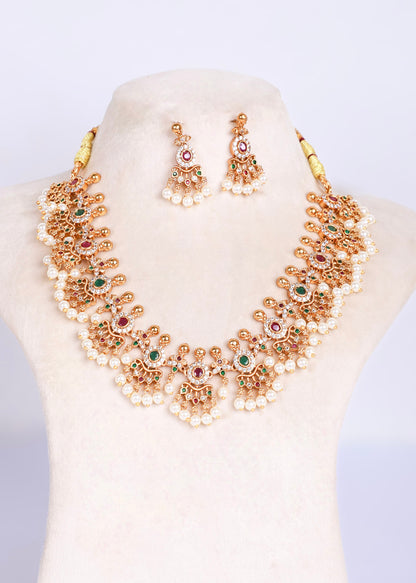 An ornate gold-toned statement necklace with matching drop earrings, displayed on a cream-colored jewelry bust. The jewelry features intricate filigree detailing, white faux pearls, and small red and green gemstone accents.