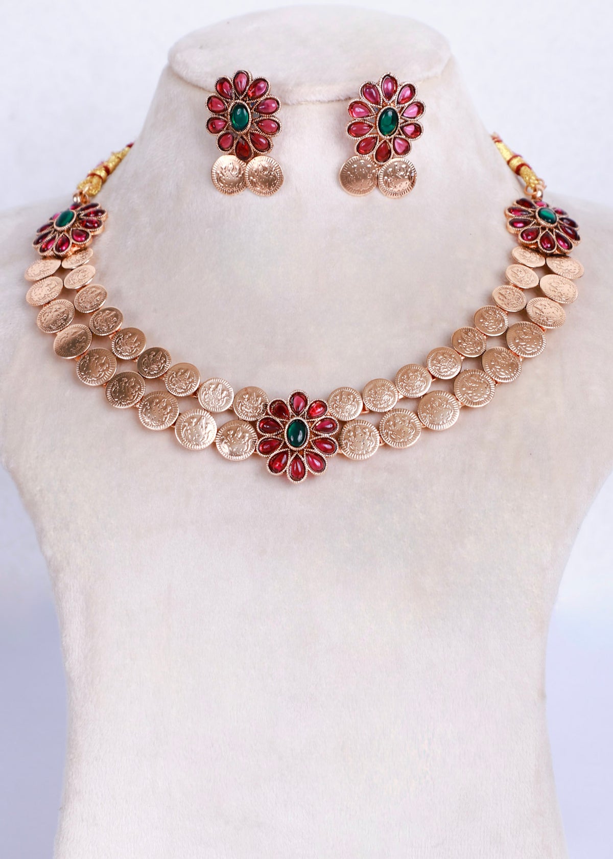 Gold coin necklace and matching stud earrings displayed on a cream jewelry bust. The necklace features a row of gold coin-like discs with three central flower motifs, each with red petal-like stones and a green center, with small gold coin drops below.