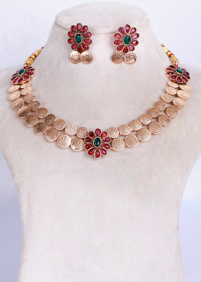 Gold coin necklace and matching stud earrings displayed on a cream jewelry bust. The necklace features a row of gold coin-like discs with three central flower motifs, each with red petal-like stones and a green center, with small gold coin drops below.