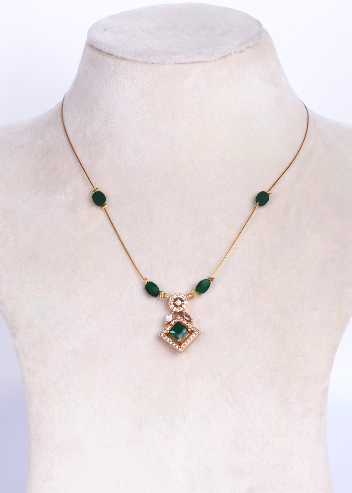 A gold chain necklace with green oval beads and a central diamond-shaped pendant, displayed on a cream-colored mannequin bust.