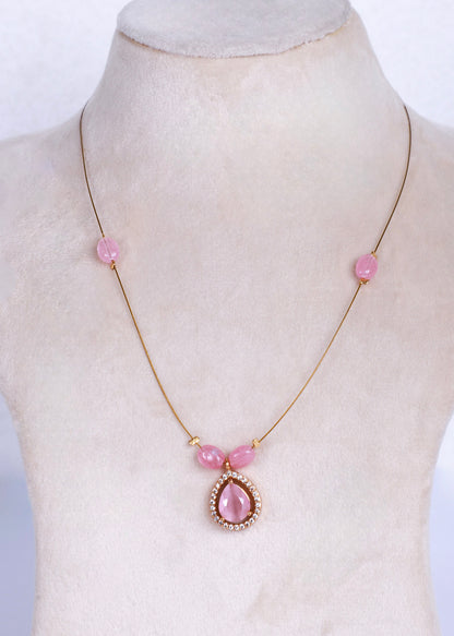 A delicate gold chain necklace displayed on a cream-colored jewelry bust. The necklace features three small pink beads spaced along the chain, with a larger teardrop-shaped pink stone pendant in a gold setting at the center.