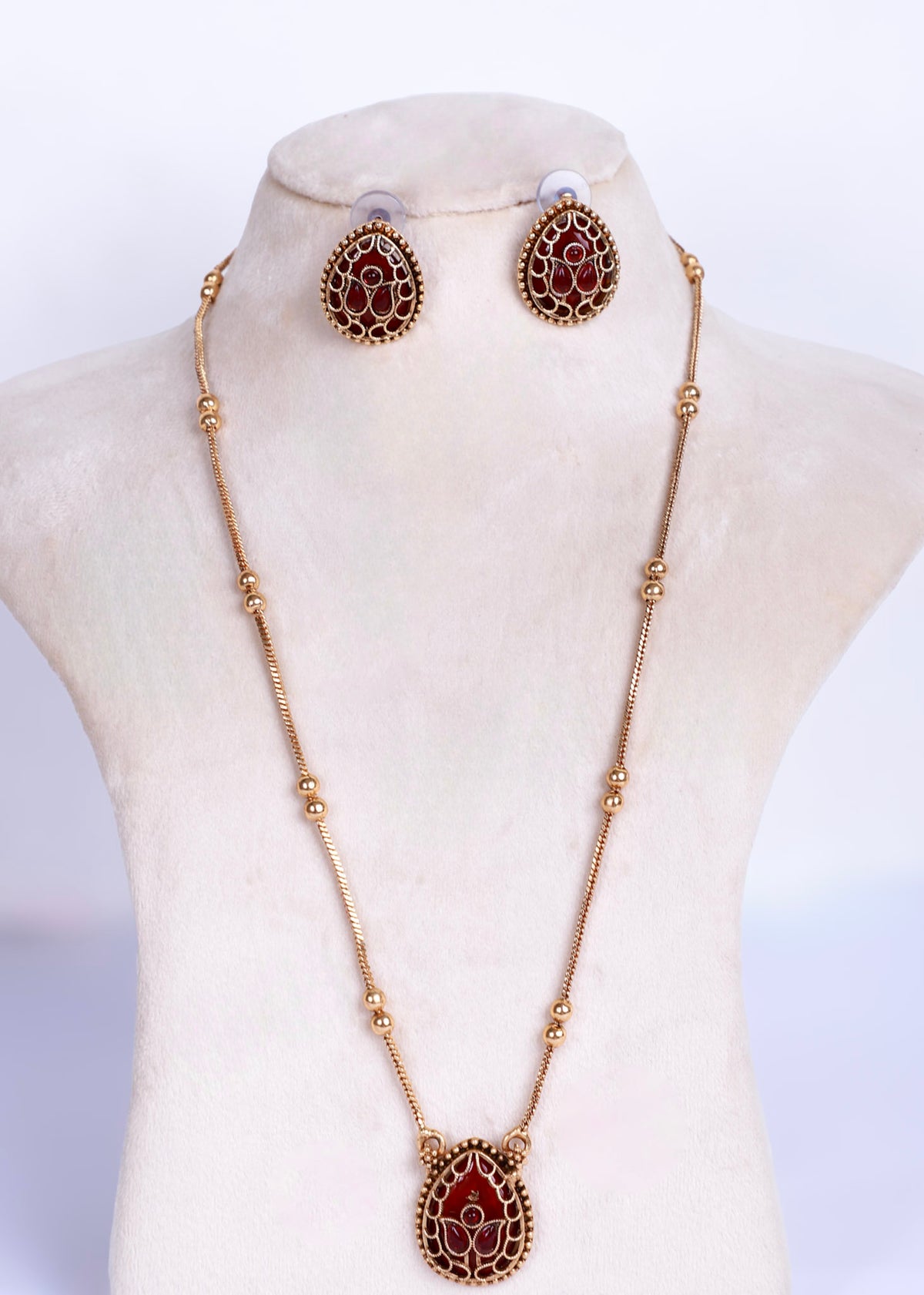 Gold-tone necklace and matching stud earrings with teardrop-shaped red stone inlays, displayed on a cream jewelry bust against a white background.