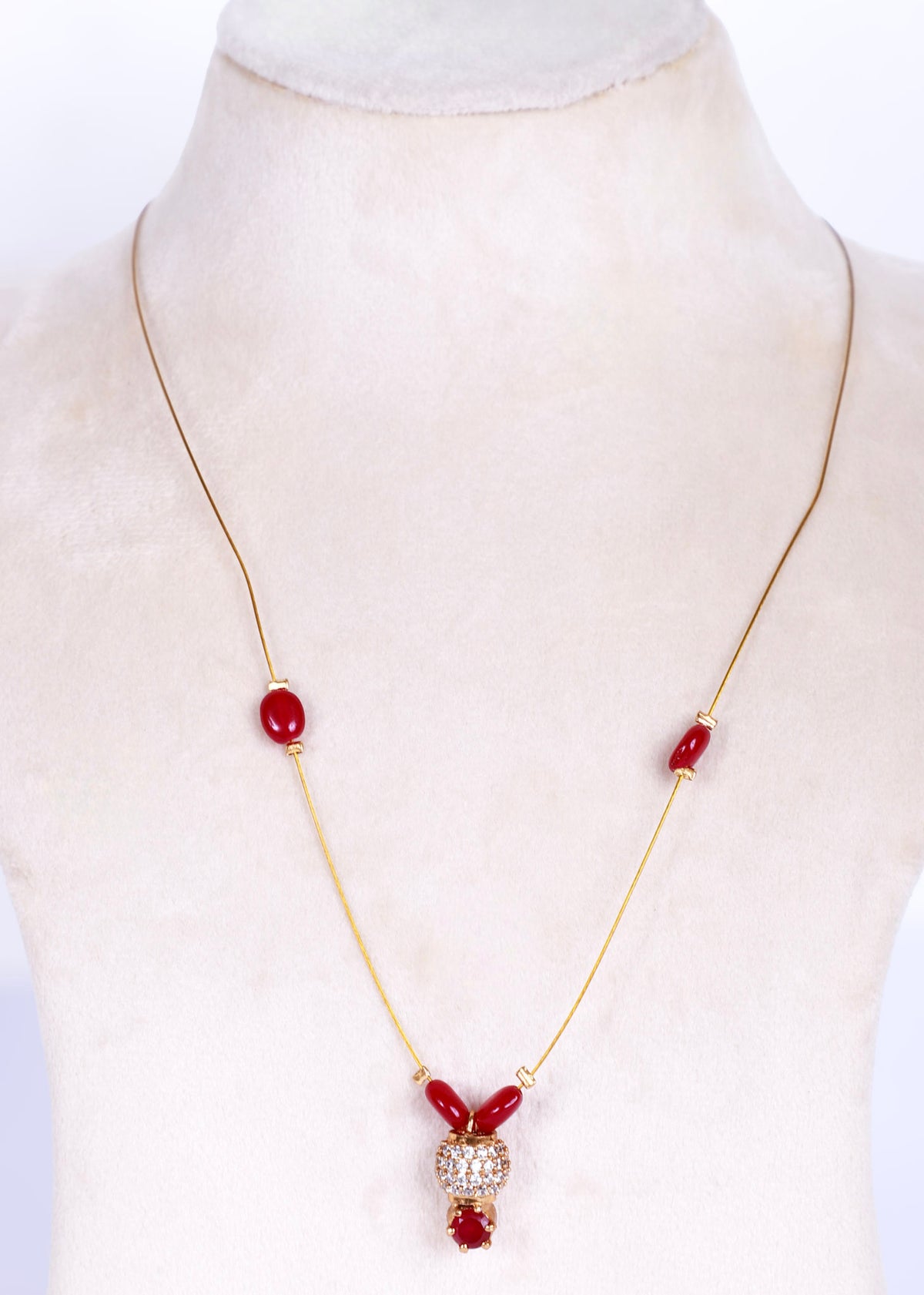 A gold chain necklace with red beads and a central crystal-encrusted pendant, displayed on a cream-colored mannequin bust.