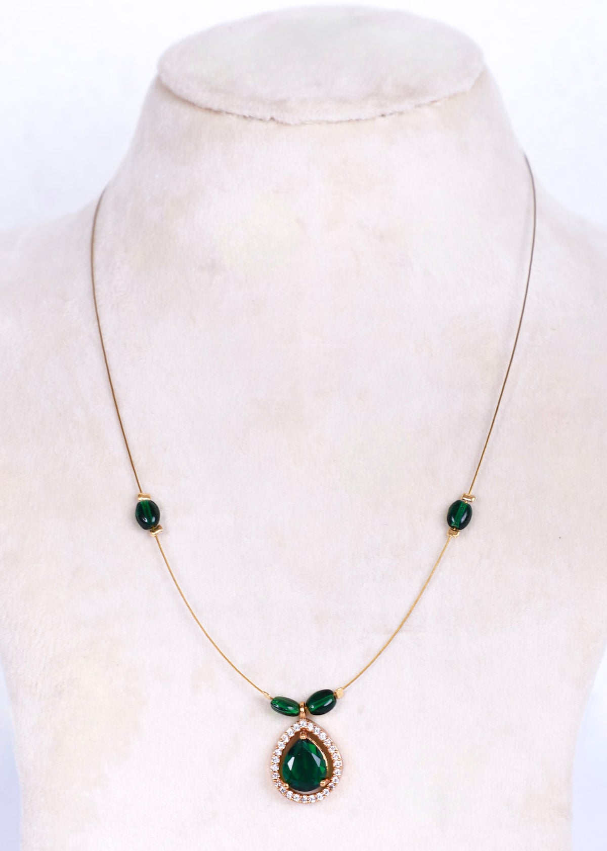 A gold chain necklace with three green gemstone accents and a teardrop-shaped green pendant surrounded by small clear stones, displayed on a cream-colored mannequin bust.