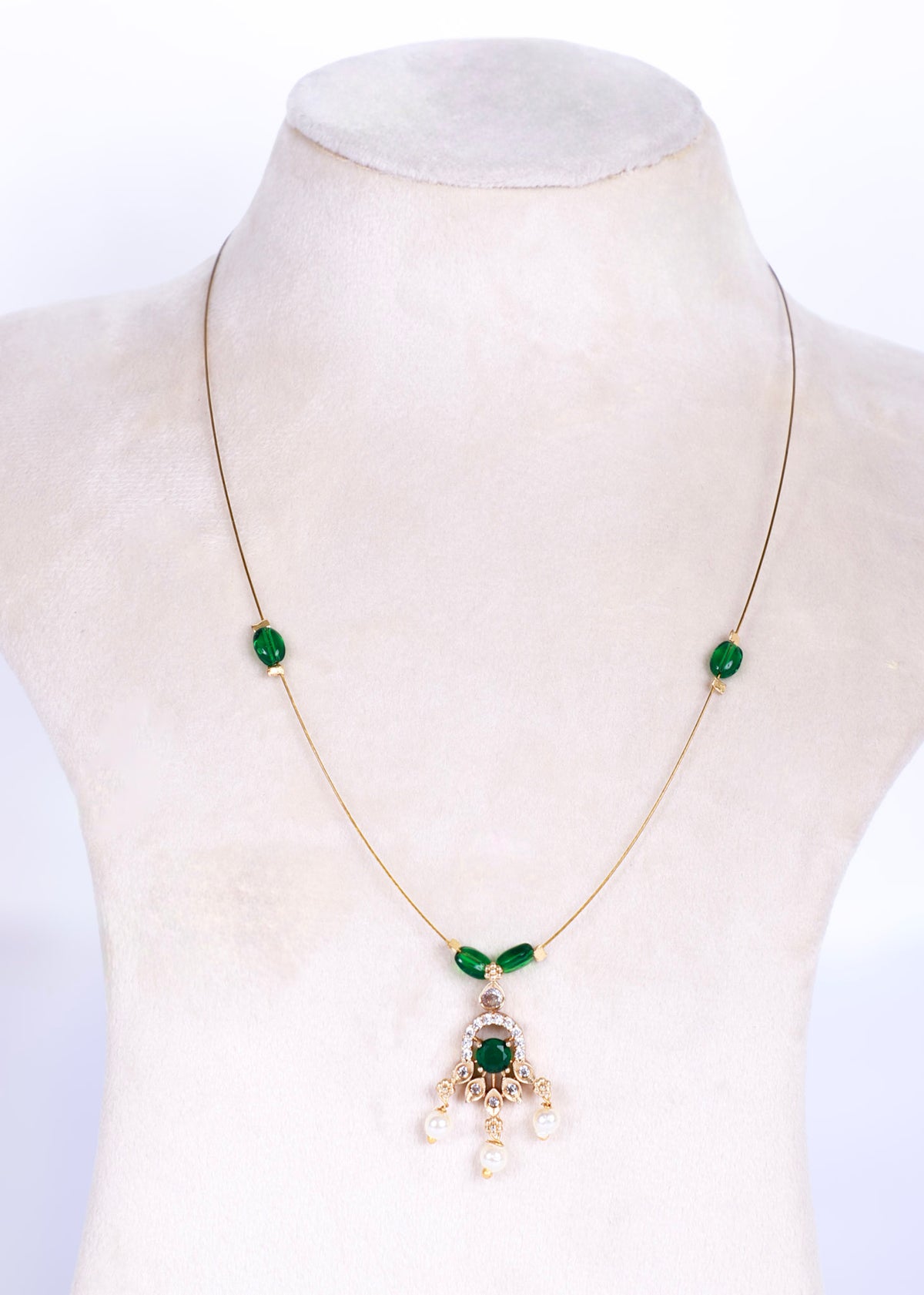 A gold chain necklace displayed on a cream-colored jewelry bust. The necklace features two small green oval stones on either side of the chest, a small green leaf-shaped accent at the center of the chain, and a central pendant with a larger green oval stone, gold detailing, and three dangling white pearl-like beads.