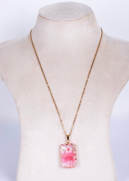 A rectangular pendant necklace with a pink and white floral pattern, suspended on anti tarnish gold-toned chain, displayed on a cream-colored mannequin bust.