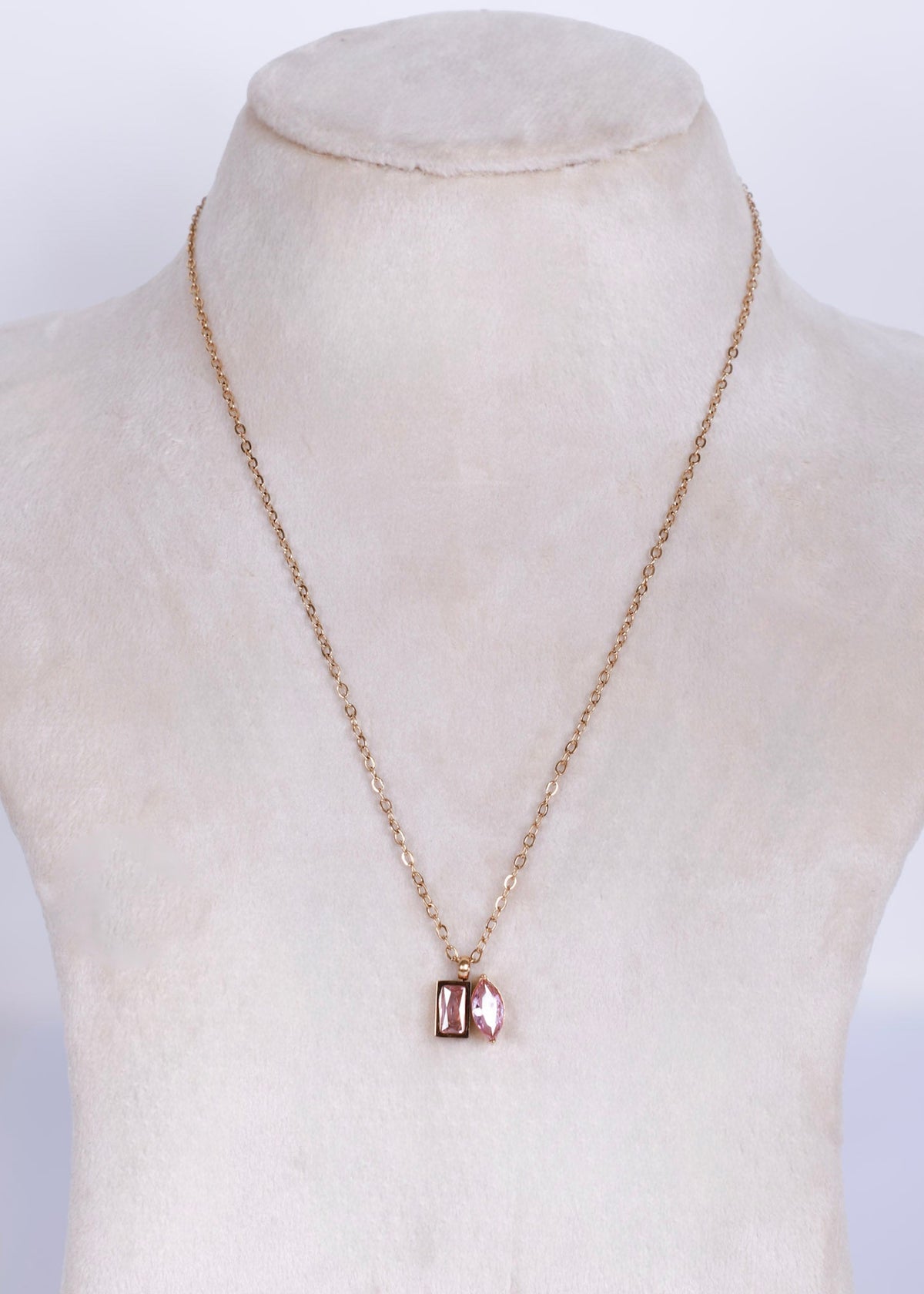 A delicate rose gold chain necklace with a small rectangular pendant featuring a pinkish-red stone, displayed on a cream-colored mannequin bust.