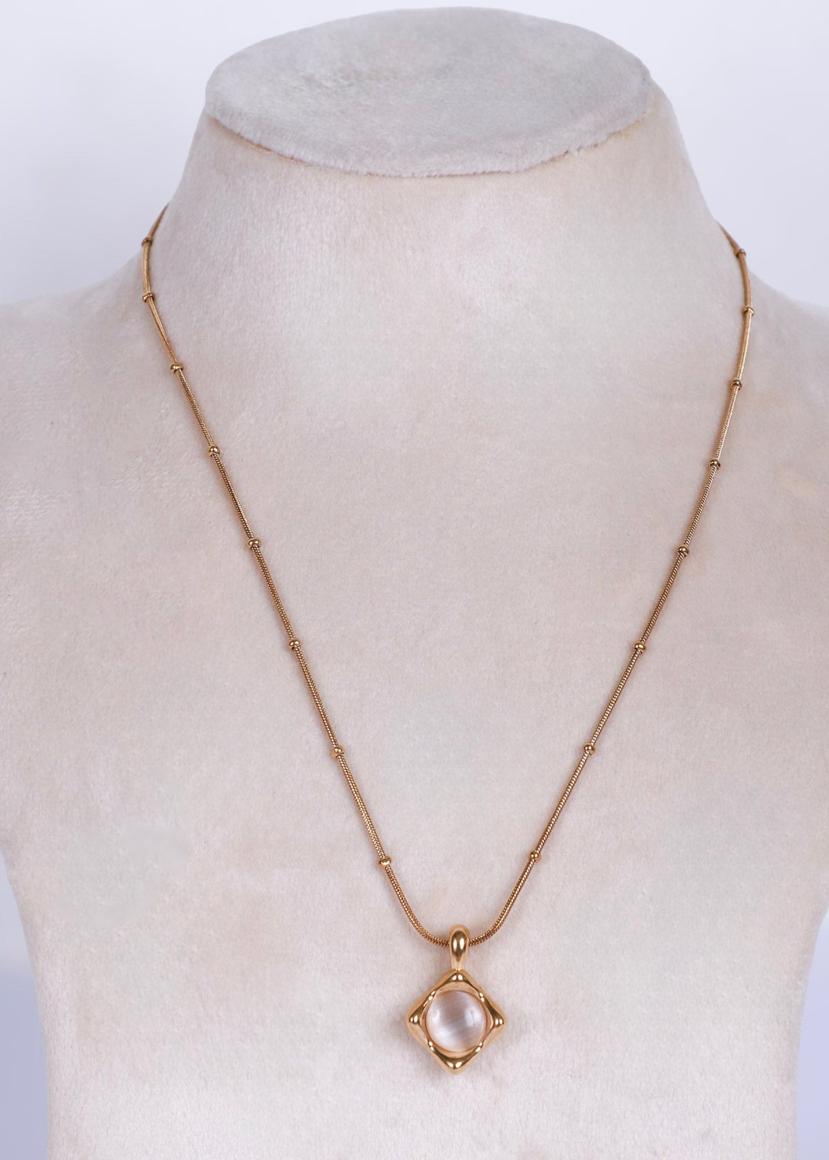 A gold chain necklace with a small oval pendant displayed on a cream-colored jewelry bust. The pendant features a pale pink stone surrounded by a gold frame.