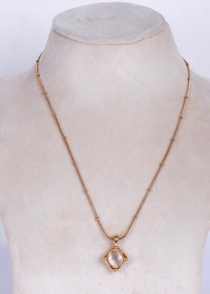 A gold chain necklace with a small oval pendant displayed on a cream-colored jewelry bust. The pendant features a pale pink stone surrounded by a gold frame.