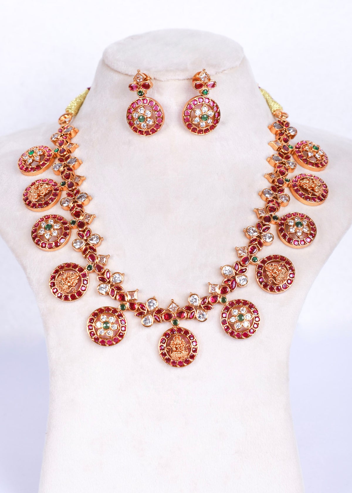 A gold-plated jewelry set featuring a statement necklace and matching drop earrings, embellished with red stones, green stones, and clear rhinestones on a cream-colored mannequin bust.