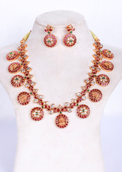 A gold-plated jewelry set featuring a statement necklace and matching drop earrings, embellished with red stones, green stones, and clear rhinestones on a cream-colored mannequin bust.
