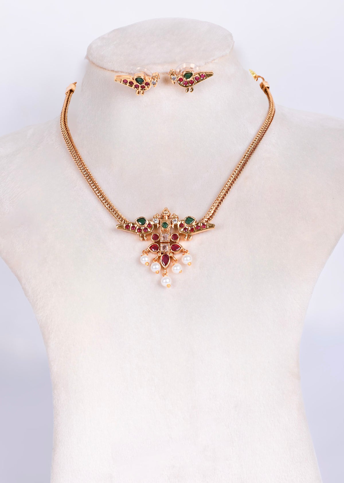 Gold-tone jewelry set displayed on a cream mannequin bust, featuring a necklace with a floral pendant, matching earrings, and a small bracelet, decorated with green and red stones and white faux pearls.