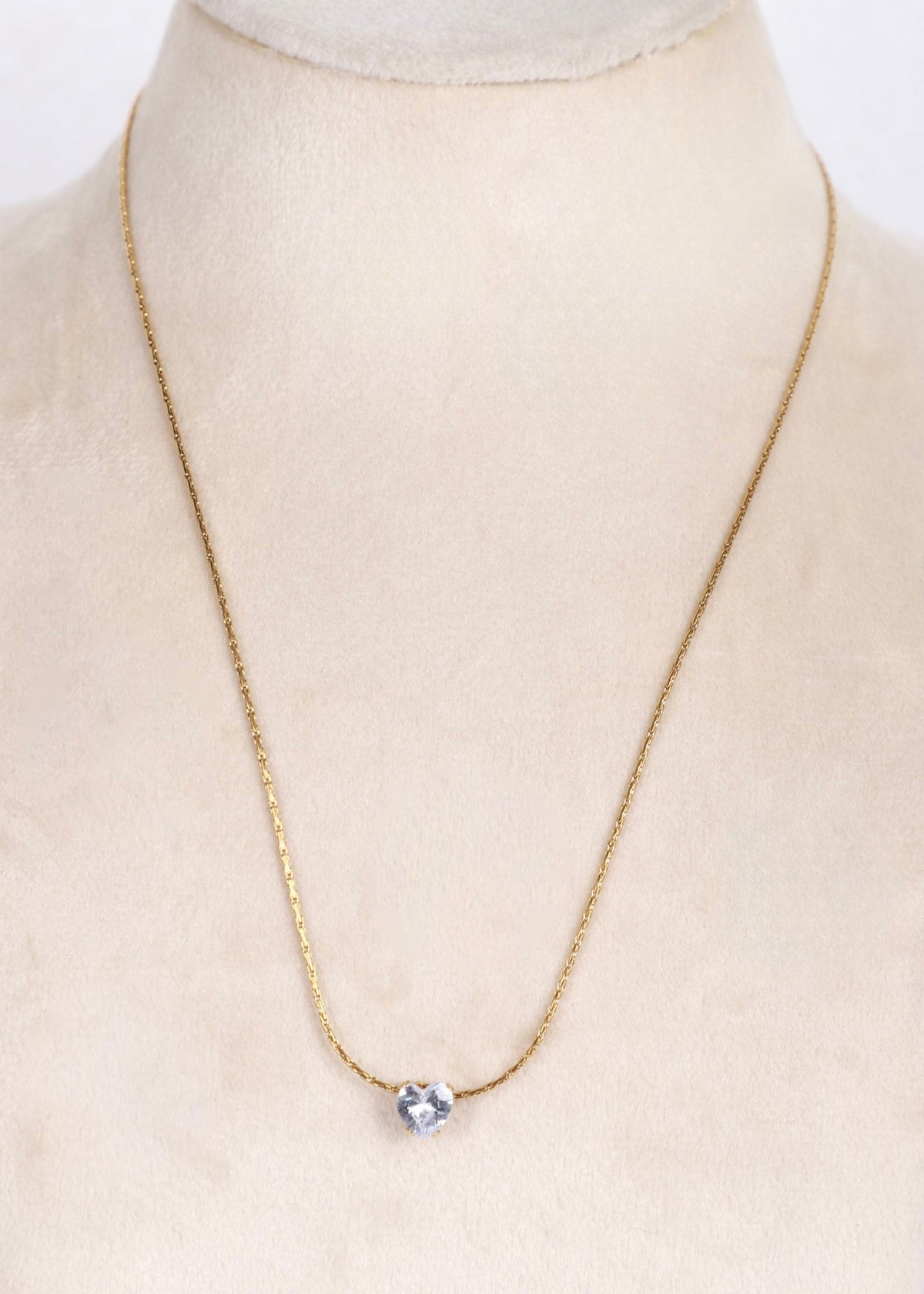 A delicate gold chain necklace with a single clear heart-shaped crystal pendant, displayed on a cream-colored jewelry bust.