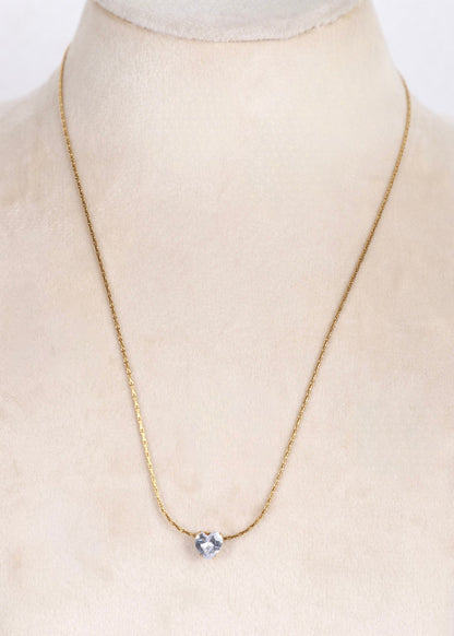 A delicate gold chain necklace with a single clear heart-shaped crystal pendant, displayed on a cream-colored jewelry bust.