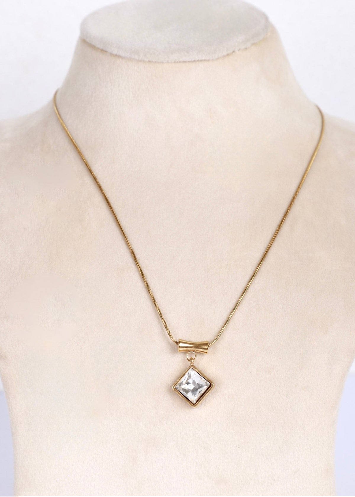 A gold chain necklace with a small diamond-shaped clear gemstone pendant, displayed on a cream-colored mannequin bust.