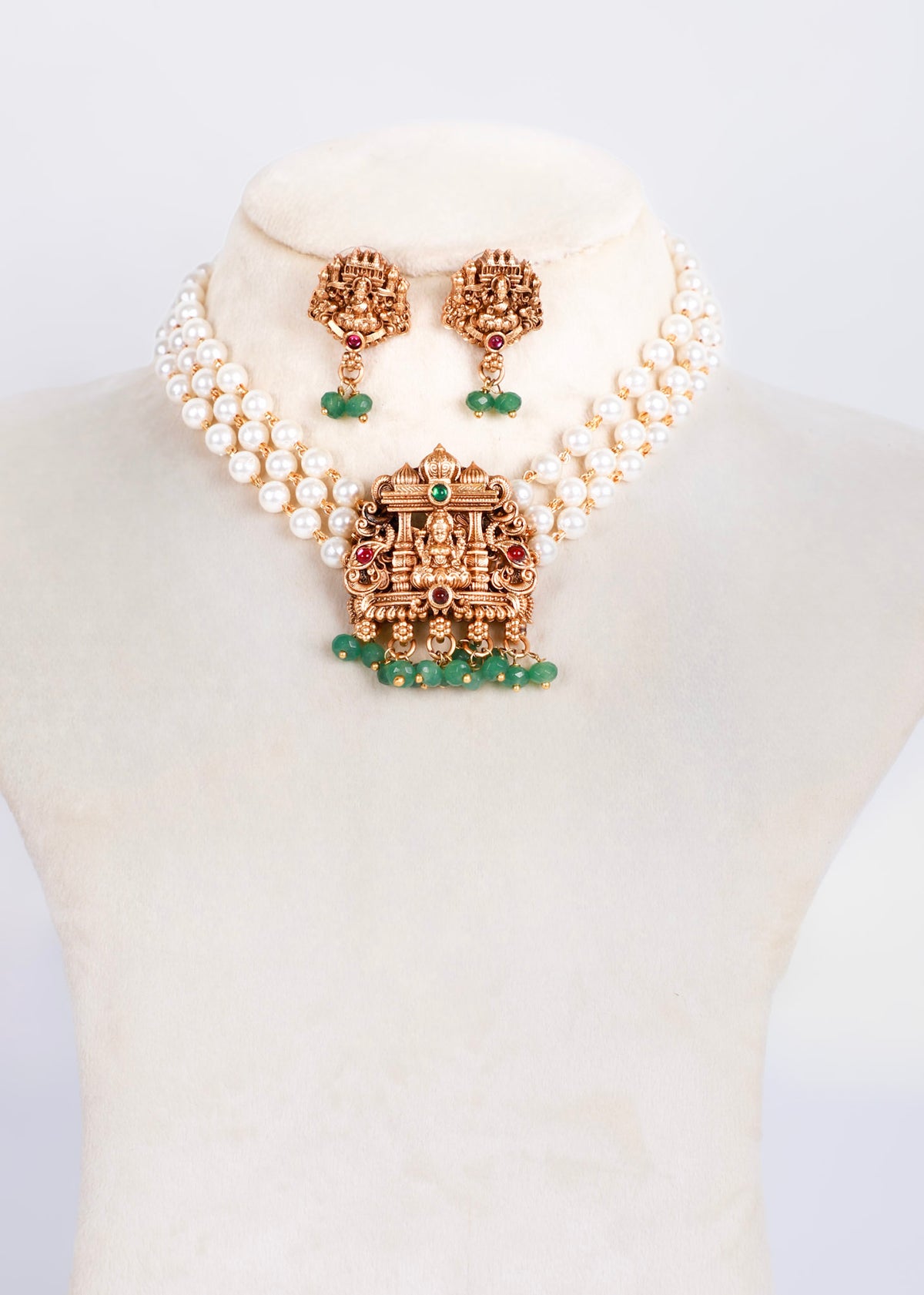 A traditional Indian jewelry set displayed on a cream-colored mannequin bust, featuring a double-strand pearl necklace with a detailed gold-toned Golu pendant, matching drop earrings, and green stone accents.