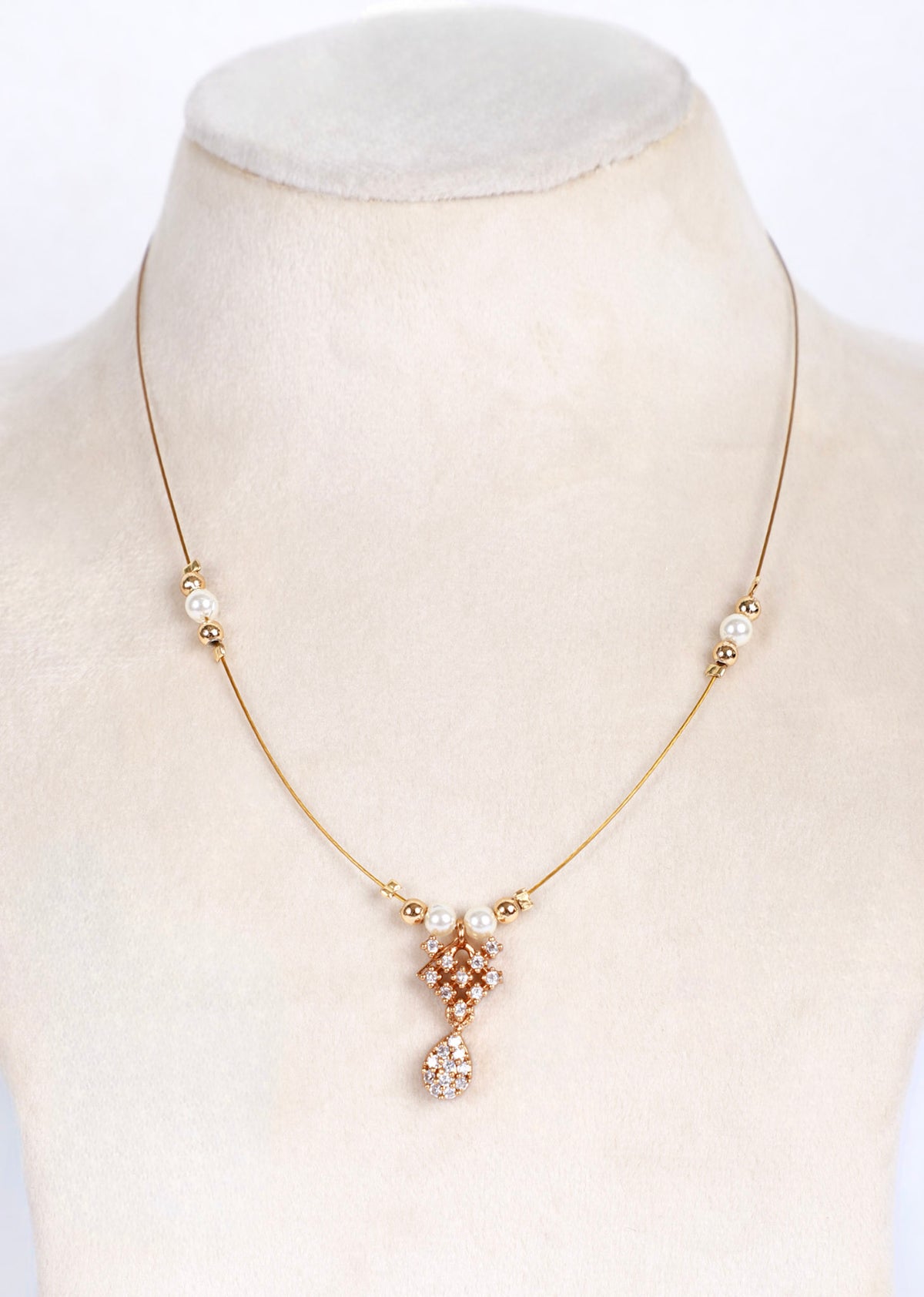 A delicate gold chain necklace displayed on a cream-colored jewelry bust. The necklace features small pearl-like beads spaced along the chain and a central pendant with a gold-toned geometric pattern accented by clear rhinestones and a dangling clear teardrop charm.
