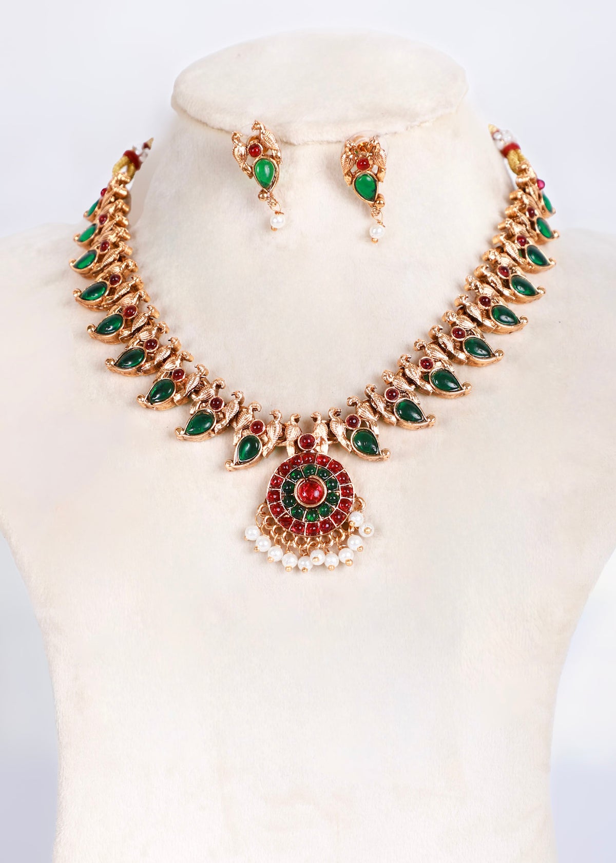 Gold-plated Indian style jewelry set featuring a necklace and matching earrings with green and red stones and white pearl accents, displayed on a cream-colored mannequin bust.