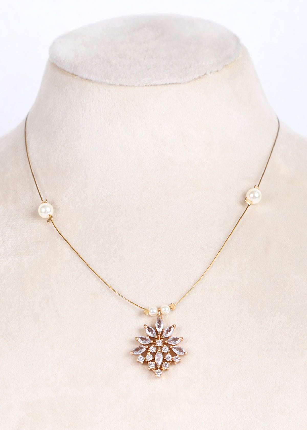 A delicate gold chain necklace displayed on a cream-colored mannequin bust. The necklace features small pearl-like beads on either side of the chain and a central gold pendant with clear rhinestones arranged in a floral or snowflake pattern.
