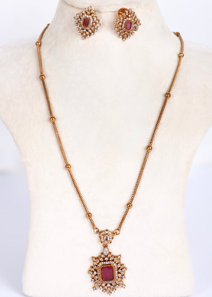 Gold-toned necklace and matching stud earrings displayed on a cream jewelry bust. The pendant and earrings feature ornate floral detailing with small clear stones and a central rectangular maroon stone.