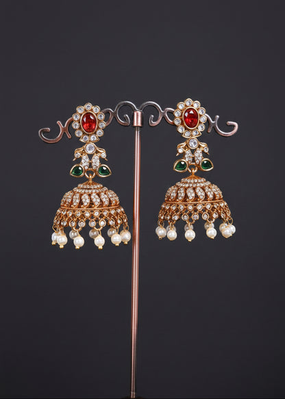 A pair of ornate gold-toned Indian jhumka earrings. Each earring features a red oval stone at the top, green stone accents, a detailed filigree dangle, and a row of small white pearl-like beads hanging at the bottom.