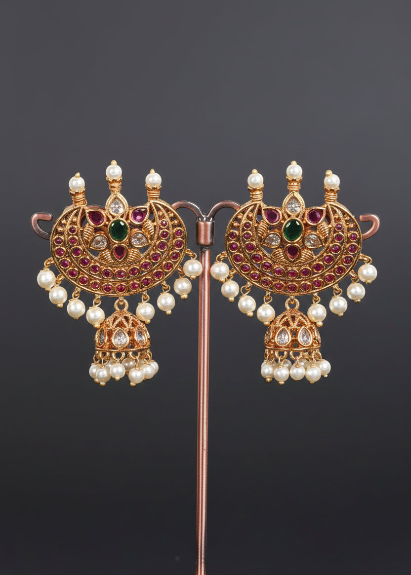 A pair of ornate gold-toned traditional earrings. Each earring features a large semi-circular pavé top with pink stones, a central green stone, white faux pearls, and a dangling jhumka with pearl beads.