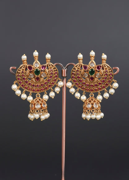 A pair of ornate gold-toned traditional earrings. Each earring features a large semi-circular pavé top with pink stones, a central green stone, white faux pearls, and a dangling jhumka with pearl beads.