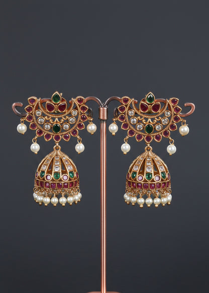 A pair of ornate gold-toned traditional Indian jhumka earrings. The earrings feature green and pink stones, clear rhinestones, and dangling white pearl bead chains.