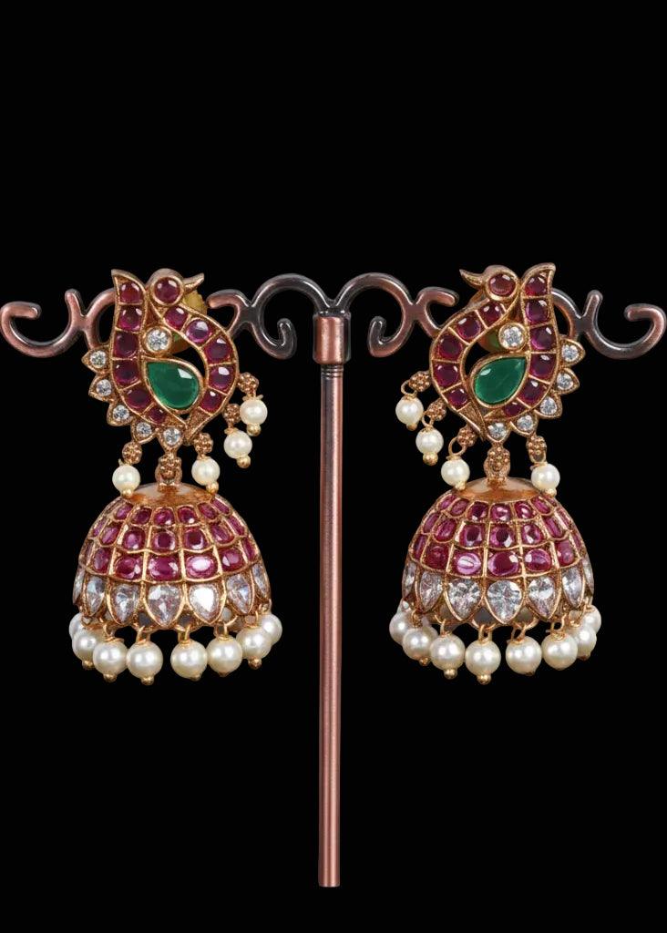 A pair of ornate Indian jhumka earrings. The earrings feature gold-plated metal frames with pink and light purple stones, a central green stone, and rows of white faux pearls hanging from the bottom.