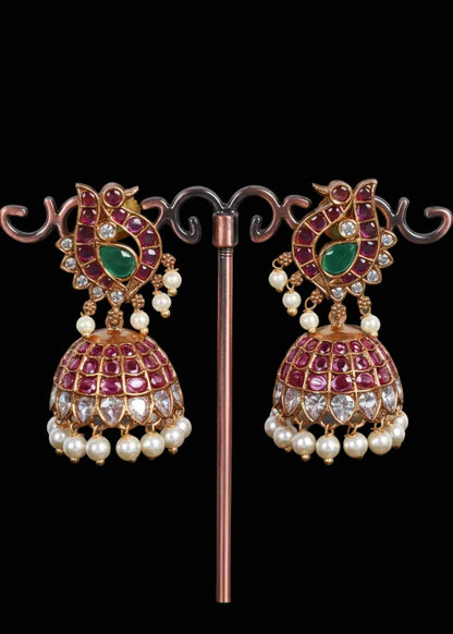 A pair of ornate Indian jhumka earrings. The earrings feature gold-plated metal frames with pink and light purple stones, a central green stone, and rows of white faux pearls hanging from the bottom.