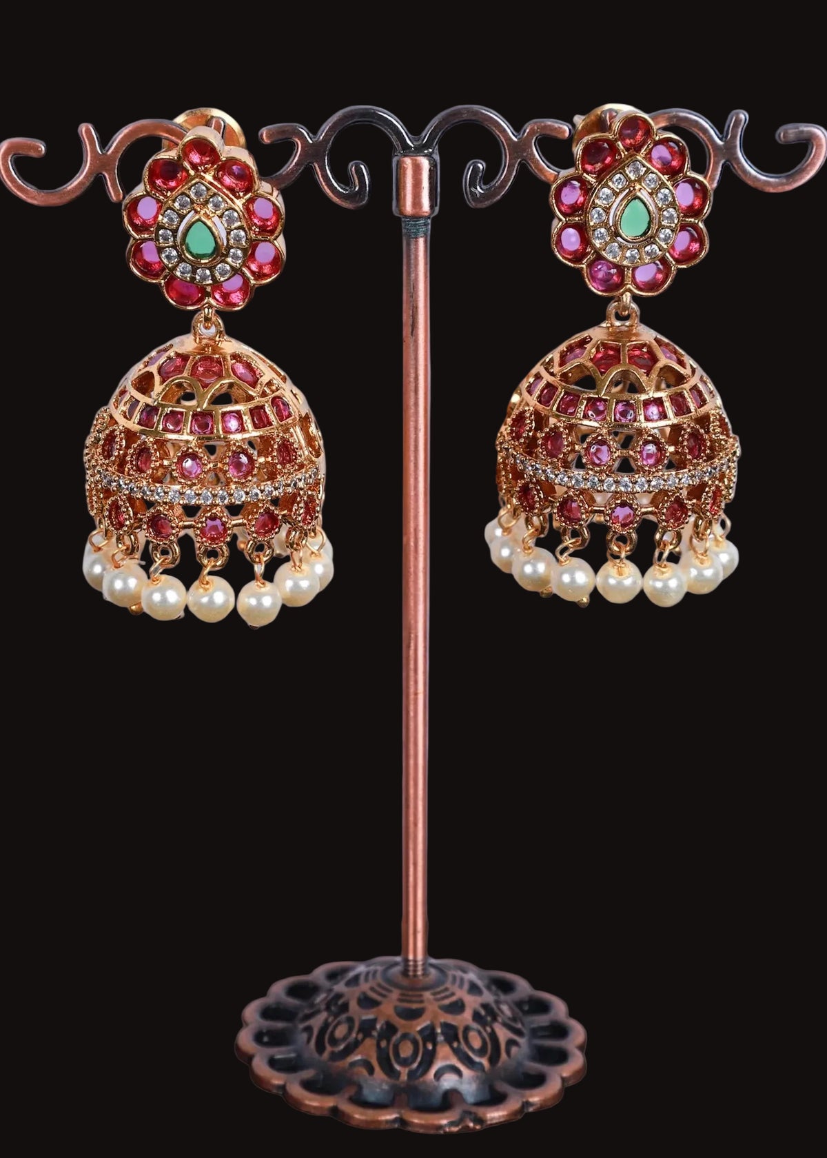 A pair of ornate Indian jhumka earrings. The earrings feature gold-toned filigree domes decorated with pink and red stones, a central green stone, and dangling white faux pearls.