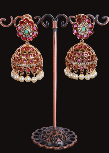 A pair of ornate Indian jhumka earrings. The earrings feature gold-toned filigree domes decorated with pink and red stones, a central green stone, and dangling white faux pearls.