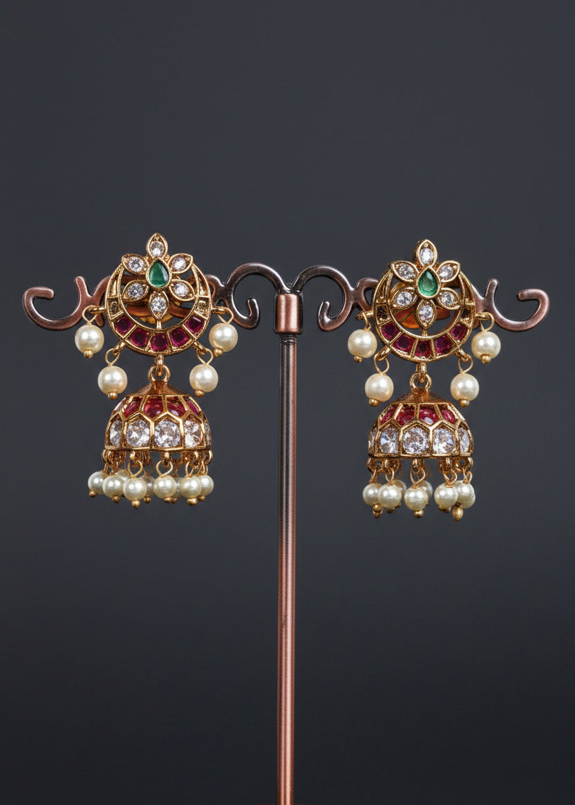 A pair of ornate gold-toned jhumka earrings with green and pink stones and white pearl drops.