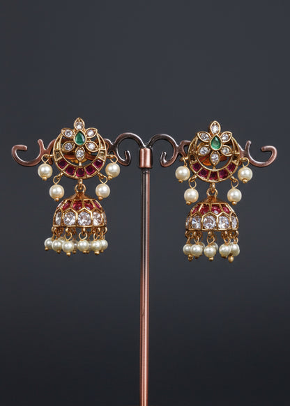 A pair of ornate gold-toned jhumka earrings with green and pink stones and white pearl drops.