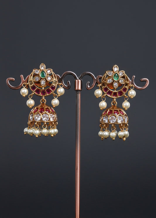 A pair of ornate gold-toned jhumka earrings with green and pink stones and white pearl drops.