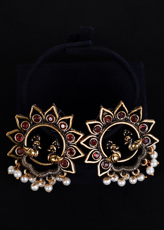 A pair of ornate gold-toned Indian jhumka earrings with peacock motifs, red stone accents, and white pearl bead drops, 