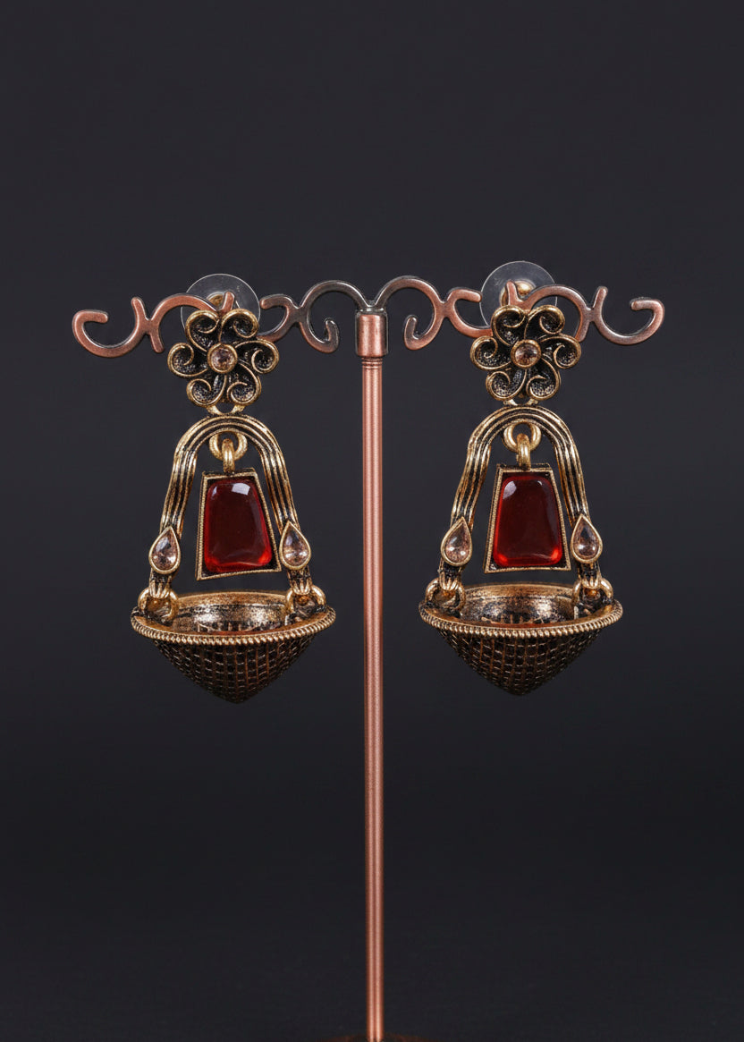 A pair of ornate dangling earrings displayed on a decorative metal stand against a light gray background. The earrings feature a bronze-toned filigree frame with a central red rectangular stone and a textured dark bowl-shaped base.