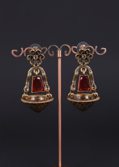 A pair of ornate dangling earrings displayed on a decorative metal stand against a light gray background. The earrings feature a bronze-toned filigree frame with a central red rectangular stone and a textured dark bowl-shaped base.