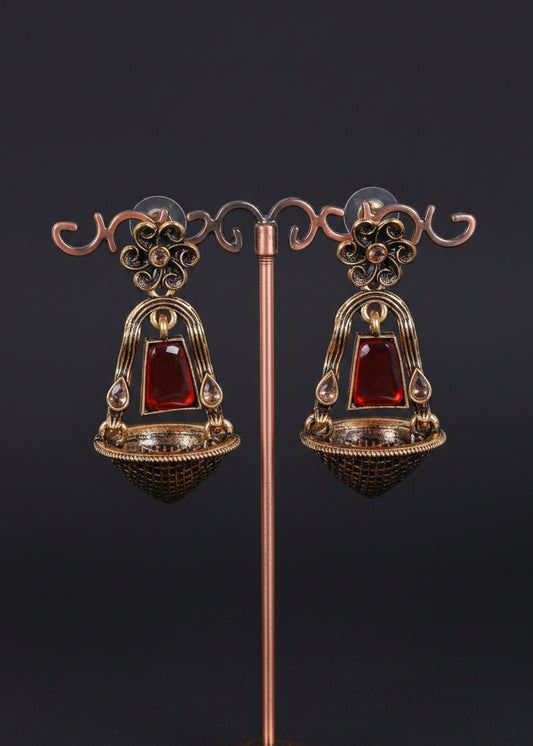 A pair of ornate dangling earrings displayed on a decorative metal stand against a light gray background. The earrings feature a bronze-toned filigree frame with a central red rectangular stone and a textured dark bowl-shaped base.
