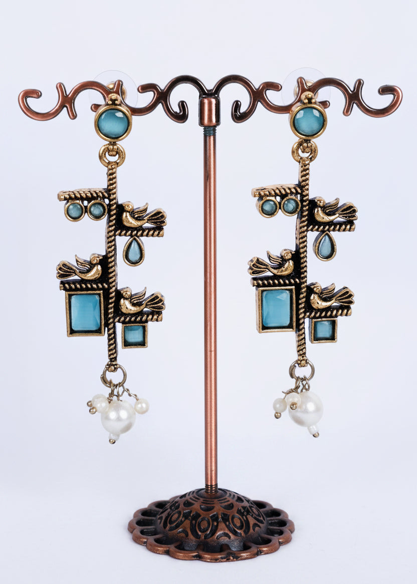 A pair of ornate dangling earrings displayed on a decorative metal stand. The earrings feature antique bronze-toned metalwork with turquoise blue stones, small bird motifs, teardrop details, and white pearl-like drops at the bottom.