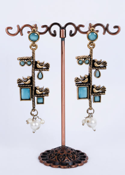 A pair of ornate dangling earrings displayed on a decorative metal stand. The earrings feature antique bronze-toned metalwork with turquoise blue stones, small bird motifs, teardrop details, and white pearl-like drops at the bottom.