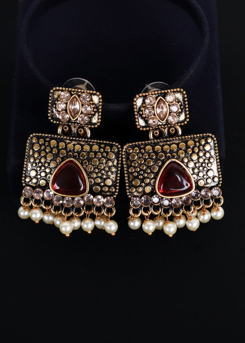 A pair of ornate Indian Kundan dangle earrings with gold-plated metalwork, red teardrop crystals, and white pearl fringe, displayed on a black velvet stand against a black background.