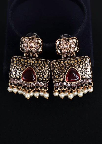 A pair of ornate Indian Kundan dangle earrings with gold-plated metalwork, red teardrop crystals, and white pearl fringe, displayed on a black velvet stand against a black background.