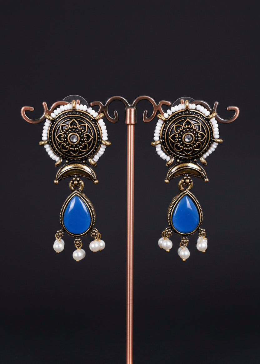 A pair of ornate Indian-style dangle earrings. The earrings feature an antique bronze circular top with intricate filigree, a small white pearl, and a prominent blue teardrop-shaped stone hanging below, finished with small white pearl drops.