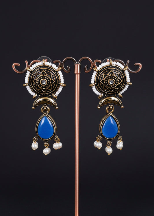 A pair of ornate Indian-style dangle earrings. The earrings feature an antique bronze circular top with intricate filigree, a small white pearl, and a prominent blue teardrop-shaped stone hanging below, finished with small white pearl drops.