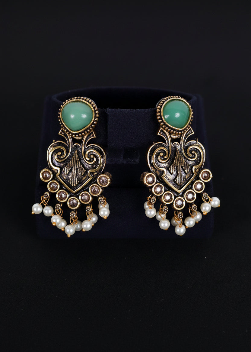 A pair of ornate gold-toned dangle earrings with mint green oval stones and white pearl drops, displayed on a black velvet jewelry stand against a black background.