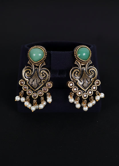 A pair of ornate gold-toned dangle earrings with mint green oval stones and white pearl drops, displayed on a black velvet jewelry stand against a black background.