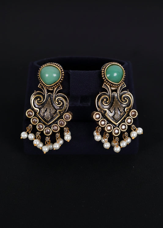 A pair of ornate gold-toned dangle earrings with mint green oval stones and white pearl drops, displayed on a black velvet jewelry stand against a black background.