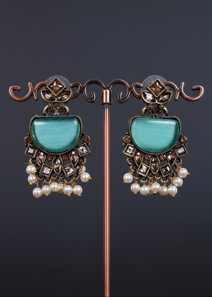 A pair of ornate dangle earrings. The earrings feature a large mint green stone in a semi-circular shape, surrounded by intricate bronze-toned metalwork, small clear rhinestones, and dangling white faux pearls.