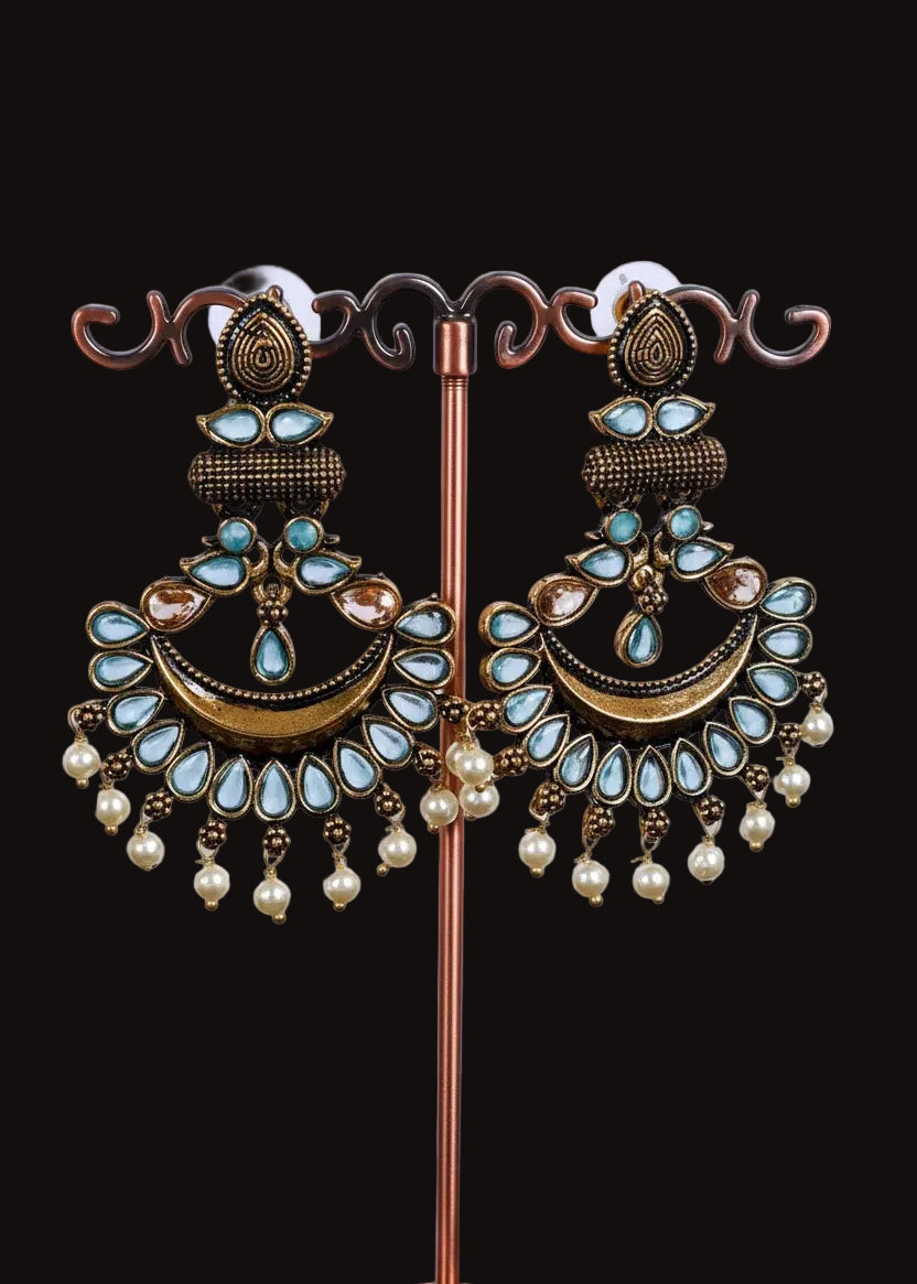 A pair of ornate gold-toned dangle earrings with light blue teardrop stones, a crescent moon design, and dangling pearl beads, displayed on a decorative stand against a light gray background.